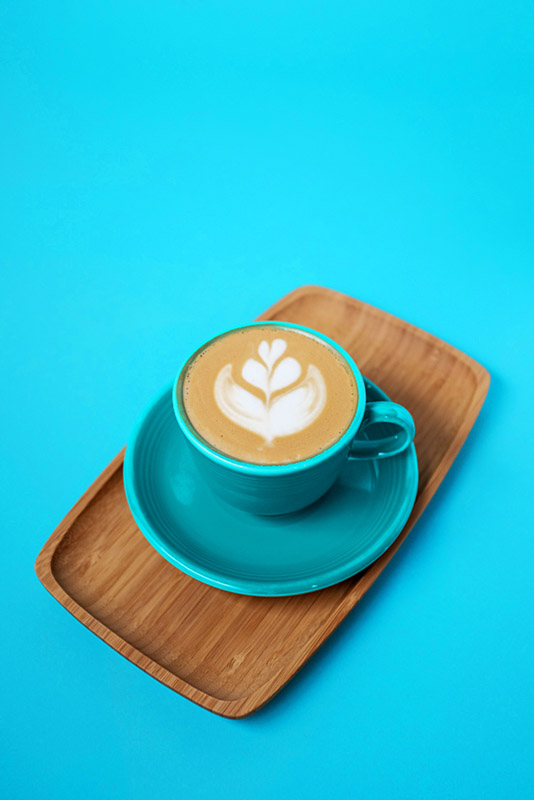 a cappuccino with latte art in a blue cup on a wooden plate against a blue backdrop
